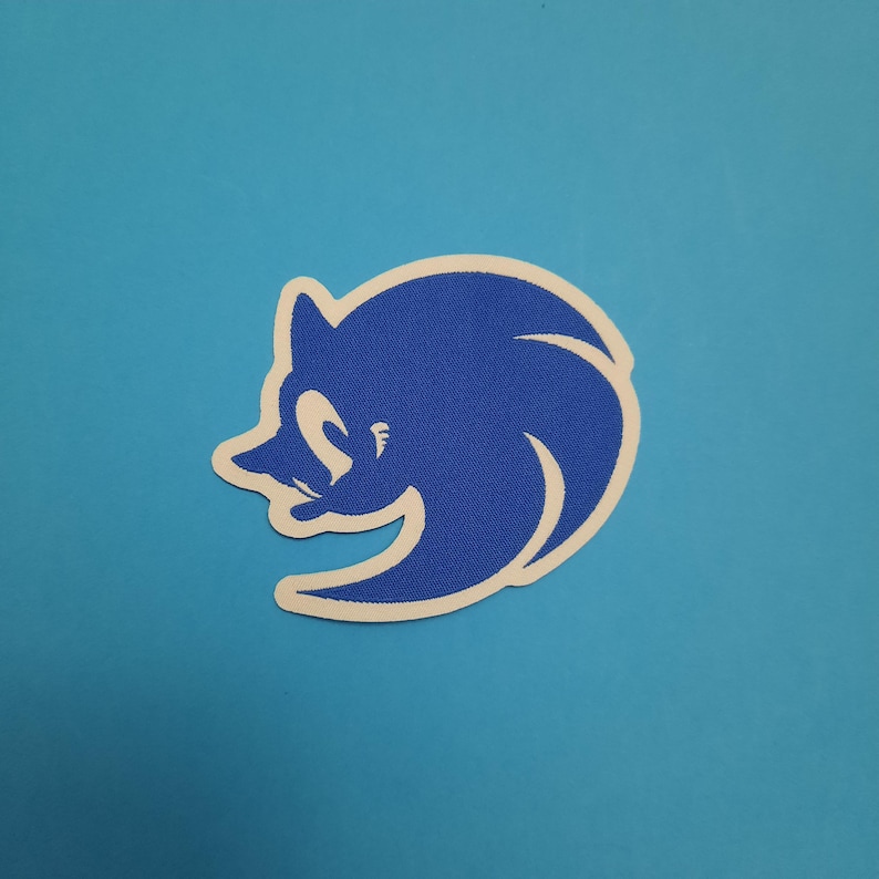 May include: A blue and white patch featuring the head of Sonic the Hedgehog. The character's face is in profile, with a smiling expression. The patch has a white border and is set against a light blue background.