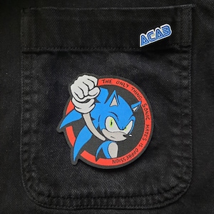 Może przedstawiać: Naszywka haftowana w kolorze czerwonym i niebieskim przedstawiająca Sonica the Hedgehog z tekstem "Jedyne, czego Sonic nienawidzi, to ucisk".