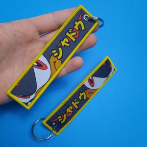 May include: Two embroidered keychains with a yellow border and Japanese text. The keychains feature a cartoon character's face in white, black, red, and orange. The background is a dark gray color.