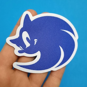 Op de afbeelding: Een blauw en wit geborduurd embleem met het iconische personage Sonic the Hedgehog. Het embleem heeft een witte rand en een gedetailleerd ontwerp van Sonics gezicht en lichaam. Het embleem is waarschijnlijk voor kleding of accessoires.