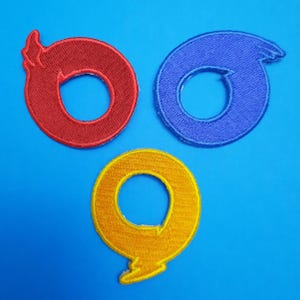 Könnte beinhalten: Drei gestickte Aufnäher in Rot, Blau und Gelb, jeweils mit einem flammenartigen Design. Die Aufnäher sind kreisförmig mit einem zentralen Loch und einer gebogenen Verlängerung. Der Hintergrund ist einfarbig blau.