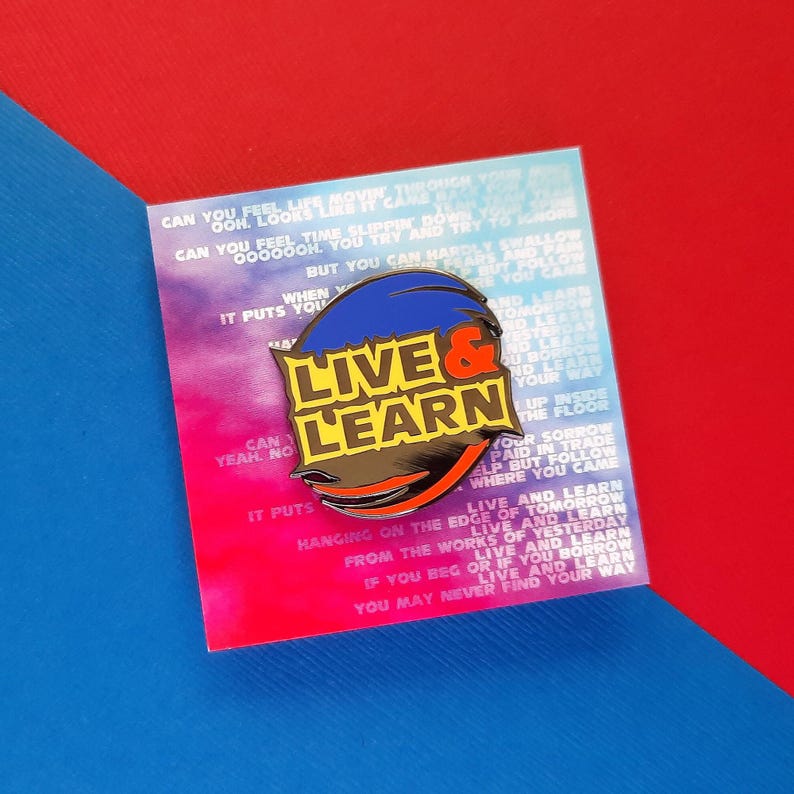 May include: A blue and yellow enamel pin with the words "Live & Learn" in a bold, white font. The pin is on a card with a colorful background and text that reads "Can you feel time slippin' down... but you can hardly swallow... it puts you hanging on the edge of tomorrow... live and learn from the works of yesterday... if you beg or if you borrow... live and learn... you may never find your way."