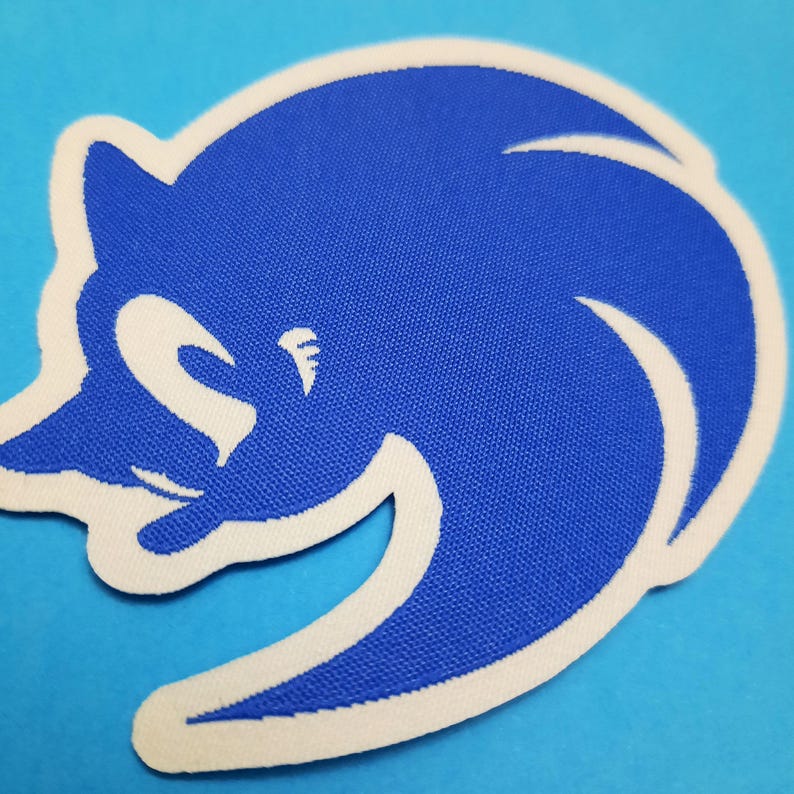 May include: A blue and white embroidered patch featuring a stylized cartoon character. The character is blue with a white outline and details. The patch is set against a light blue background.
