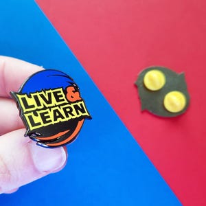 May include: A blue, black, orange, and yellow enamel pin with the words "Live & Learn" in a bold, white font.