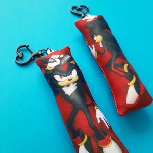 May include: Two red and black keychains featuring a cartoon character. The keychains have a heart-shaped clasp. The character is black with red accents, a white chest, and yellow details. The background is a solid blue.