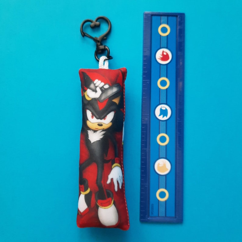 May include: A red and black rectangular keychain featuring a cartoon character. The keychain has a black heart-shaped clasp. A blue ruler with white and yellow circles and symbols is next to the keychain.