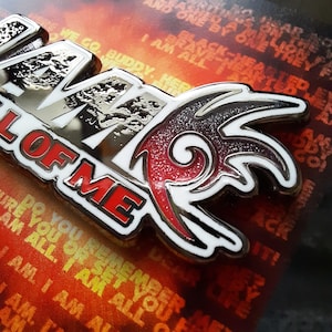 May include: A silver and red enamel pin with the words "All Of Me" and a stylized flame design. The pin is on a background of orange and yellow.