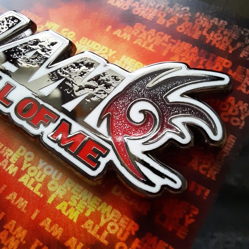 May include: A silver and red enamel pin with the words "All Of Me" and a stylized flame design. The pin is on a background of orange and yellow.