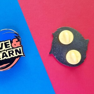 May include: A blue and black enamel pin with the text "Live & Learn" on a white background. The back of the pin is black with two yellow metal pin backs.