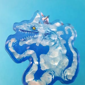 May include: A blue and white acrylic keychain featuring a cartoon monster with sharp teeth and a yellow eye. The keychain has a silver ring for attaching to a bag or keys.