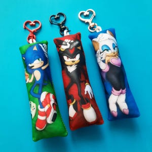 May include: Three keychains featuring characters from the Sonic the Hedgehog franchise. Each keychain has a different character printed on a rectangular pillow-shaped design. The keychains have heart-shaped clasps in red, black, and gold.