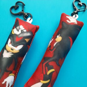 May include: Two keychains featuring a cartoon character with black, red, and yellow accents. The keychains have a heart-shaped clasp and a red and black patterned design. The background is a solid light blue.