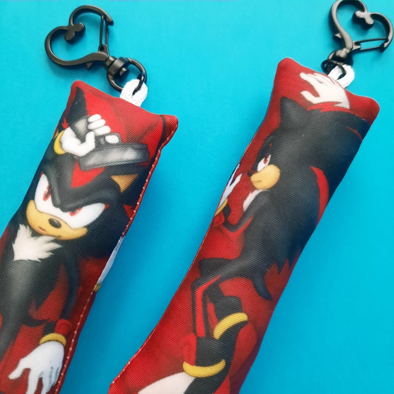 May include: Two keychains featuring a cartoon character with black, red, and yellow accents. The keychains have a heart-shaped clasp and a red and black patterned design. The background is a solid light blue.