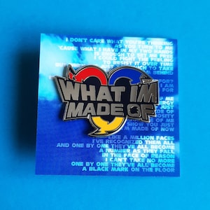 May include: A silver pin with the words "What I'm Made Of" in black letters on a blue, red, and yellow background. The pin is on a blue background with text that reads "Don't care what you're thinking as you turn to me 'cause what I have in my two hands is enough to set me free I could fight the feeling to resist it over time just which to take behind for? I am for f me ergy just de of e of me show you just I'm made of now like a million faces I've recognized them all and one by one they've all become a number as they fall in the face of reason I can't take no more one by one they've all become a black mark on the floor"