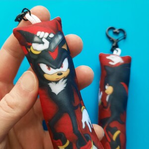 May include: Two rectangular keychains featuring a cartoon character with black, red, yellow, and white colors. The character is depicted on a red background. One keychain is held by a hand, and the other is hanging on a heart-shaped clasp.