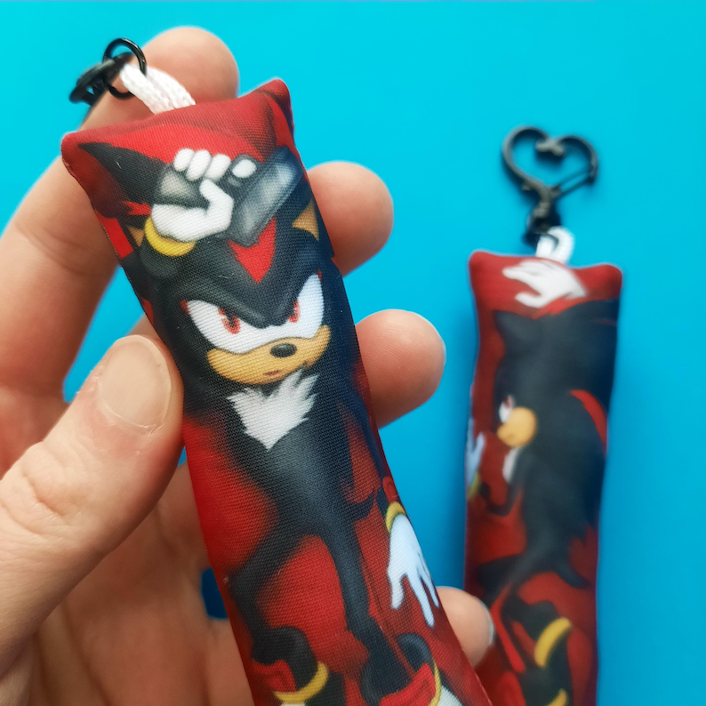 May include: Two rectangular keychains featuring a cartoon character with black, red, yellow, and white colors. The character is depicted on a red background. One keychain is held by a hand, and the other is hanging on a heart-shaped clasp.