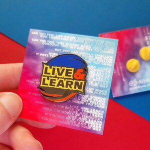 May include: A colorful enamel pin with the words "Live & Learn" in black letters on a yellow background. The pin is surrounded by a blue, red, and orange swirl. The pin is on a white card with text that reads "Can you feel her movie through you...Live and learn tomorrow and learn yesterday and learn if you beg or if you borrow live and learn you may never find your way."