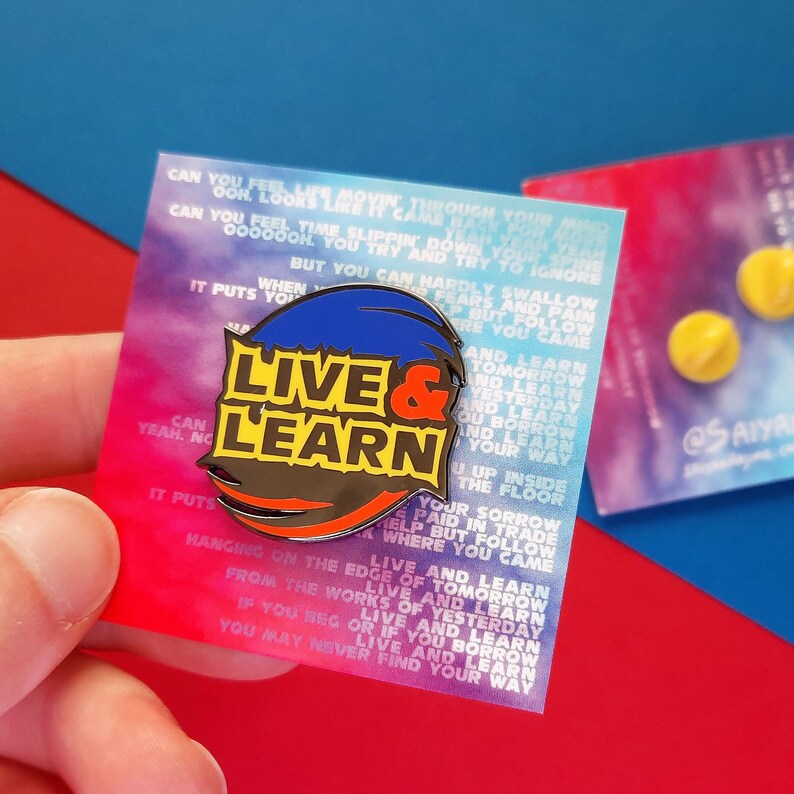 May include: A colorful enamel pin with the words "Live & Learn" in black letters on a yellow background. The pin is surrounded by a blue, red, and orange swirl. The pin is on a white card with text that reads "Can you feel her movie through you...Live and learn tomorrow and learn yesterday and learn if you beg or if you borrow live and learn you may never find your way."