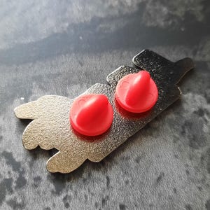 May include: A silver metal pin with two red plastic push pins. The pin features a design of a hand with two fingers pointing up.