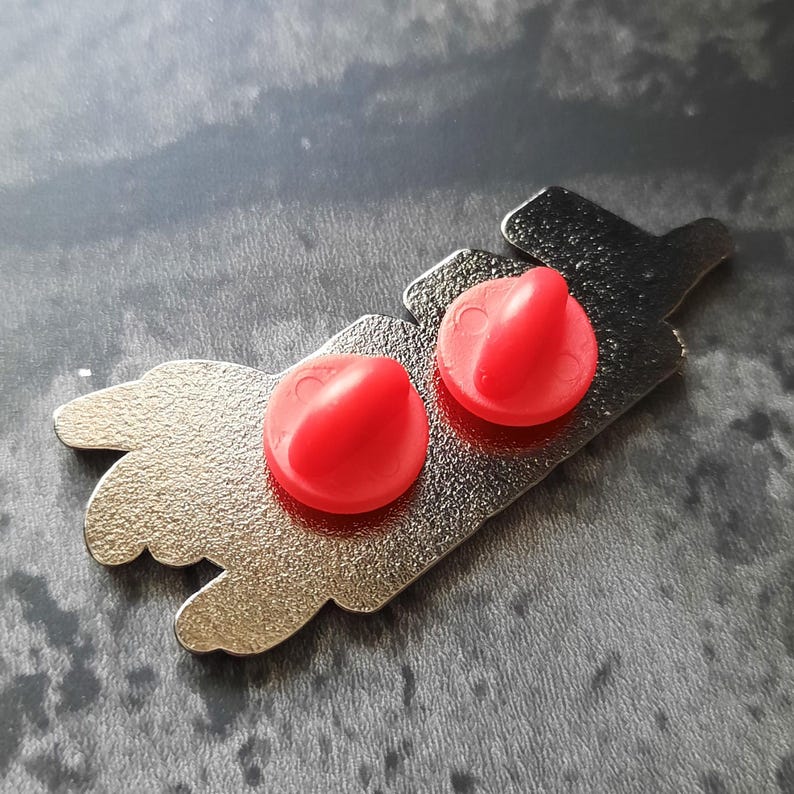 May include: A silver metal pin with two red plastic push pins. The pin features a design of a hand with two fingers pointing up.
