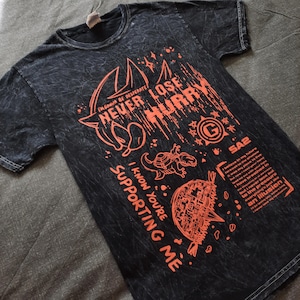May include: Black t-shirt with orange graphics and text. The design includes the phrases "NEVER LOSE HURRY" and "I KNOW YOU'RE SUPPORTING ME", along with illustrations of a turtle and other shapes. The shirt has a textured, faded appearance.