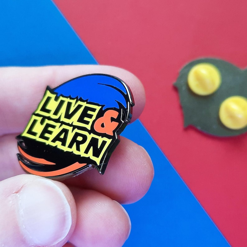 May include: A black and orange enamel pin with the words "Live & Learn" in yellow letters. The pin is shaped like a wave and has a blue and black design.