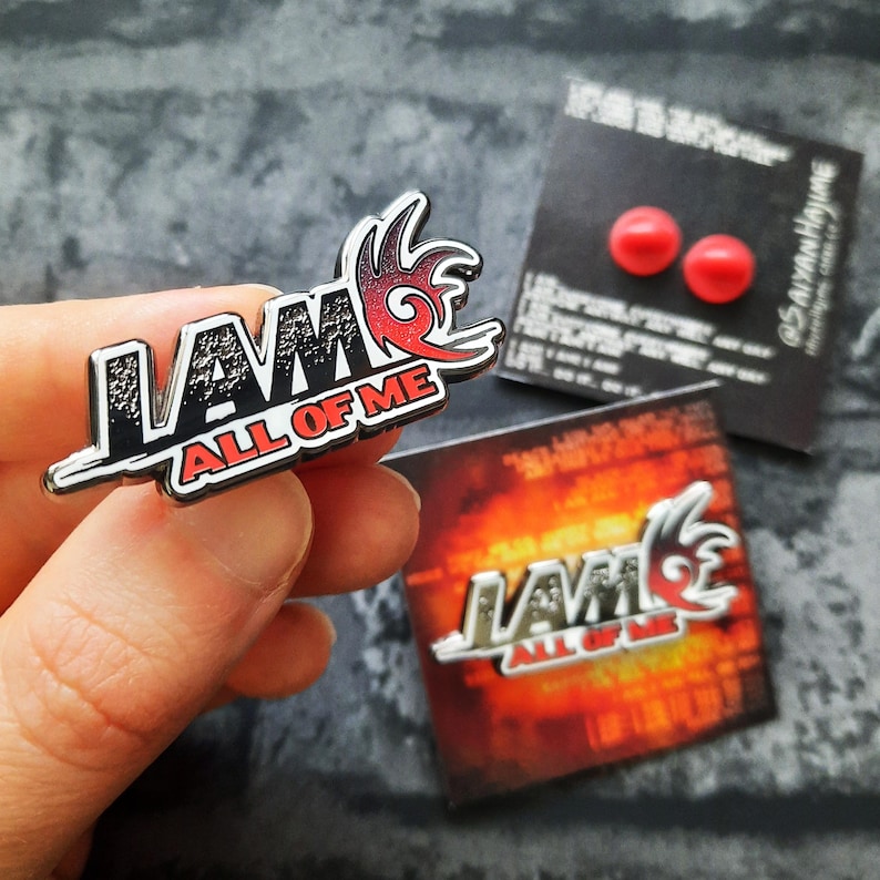 May include: A silver metal pin with a black and red design. The pin says "I AM ALL OF ME" in a bold font. The design includes a stylized flame shape.