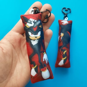 May include: Two red rectangular keychains featuring the character Shadow the Hedgehog from the Sonic the Hedgehog franchise. The keychains have black hardware and a white cord loop. The character is depicted in a dynamic pose against a red background.