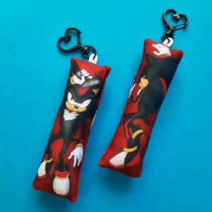 May include: Two red rectangular keyrings featuring the character Shadow the Hedgehog from the Sonic the Hedgehog franchise. Each keyring has a black heart-shaped clasp. The character is depicted in a dynamic pose against a red background.