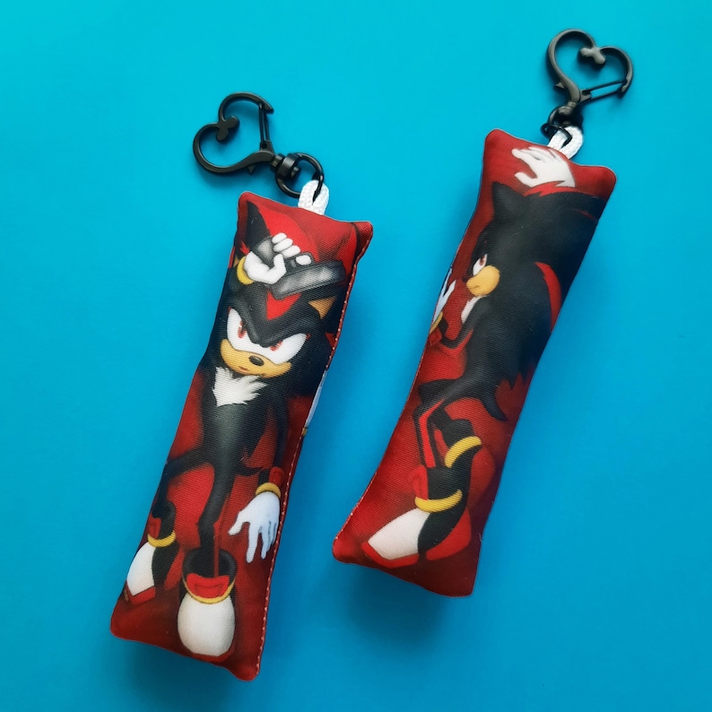 May include: Two red rectangular keychains featuring the character Shadow the Hedgehog from the Sonic the Hedgehog franchise. Each keychain has a black heart-shaped clasp. The character is depicted in a dynamic pose against a red background.