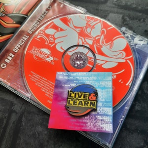 May include: A red CD case with a Sonic Adventure 2 soundtrack disc inside. The disc features a white cartoon image of Sonic the Hedgehog. A black and yellow enamel pin with the words "Live & Learn" is in the foreground.