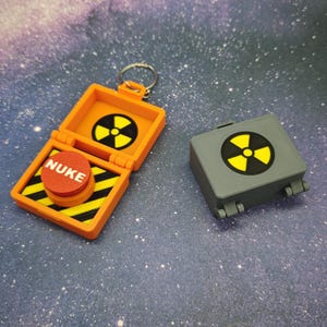 May include: Two small, plastic toy boxes with a nuclear symbol and the word "NUKE" on a red button. The boxes are orange and grey.