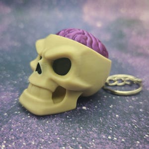 May include: A beige skull figurine with a purple brain exposed. The skull has black eye sockets and a detailed jawline. A silver ring with a braided design is next to the skull. The background is a sparkly purple.