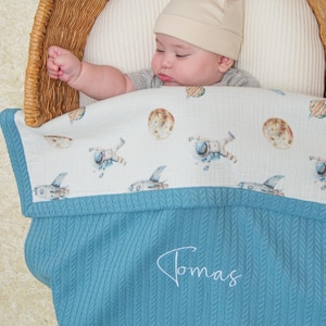 Op de afbeelding: Een baby met een beige mutsje ligt in een gevlochten rieten mand, bedekt met een blauwe deken met visgraatmotief en de naam Tomas in wit geborduurd. De deken heeft een witte bovenrand met illustraties in een ruimtevaartthema.
