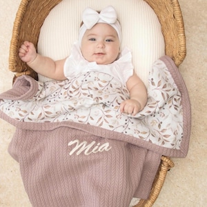 Könnte beinhalten: Eine mauvefarbene Strickdecke für Babys mit dem in weißer Schreibschrift gestickten Namen Mia, kombiniert mit einer weißen Decke mit Blumenmuster. Die Decke liegt über einem Baby mit weißem Oberteil und weißem Schleifen-Stirnband im Weidenkorb.