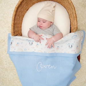 Può includere: Una coperta azzurra a nido d'ape con il nome Owen ricamato in corsivo bianco, sovrapposta a una coperta bianca con motivo di arcobaleni e nuvole. La coperta è in una cesta di vimini con un bambino che indossa un berretto beige con orecchie.
