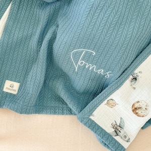 Puede incluir: Una manta de bebé de punto azul turquesa con un borde blanco y un estampado de temática espacial. La manta está personalizada con el nombre "Thomas" bordado en blanco.