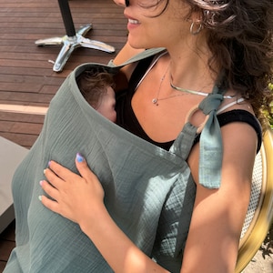 Puede incluir: Una mujer con gafas de sol y un portabeb&eacute;s de muselina verde con un beb&eacute; dentro. El portabeb&eacute;s tiene un anillo de madera y una corbata en la parte superior.