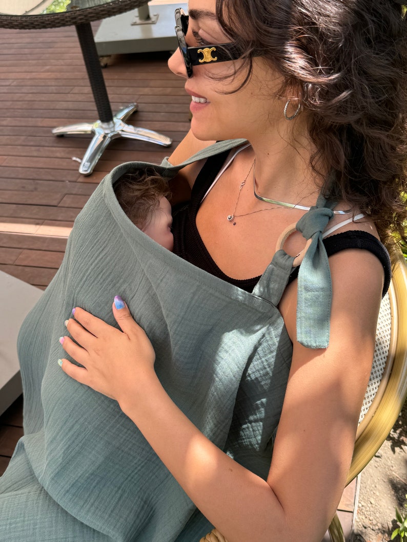 Puede incluir: Una mujer con gafas de sol y un portabeb&eacute;s de muselina verde con un beb&eacute; dentro. El portabeb&eacute;s tiene un anillo de madera y una corbata en la parte superior.