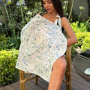 Puede incluir: Una cubierta de lactancia blanca con un patr&oacute;n de hojas verdes y azules. La cubierta tiene un anillo de madera en la parte superior para una f&aacute;cil apertura y cierre.