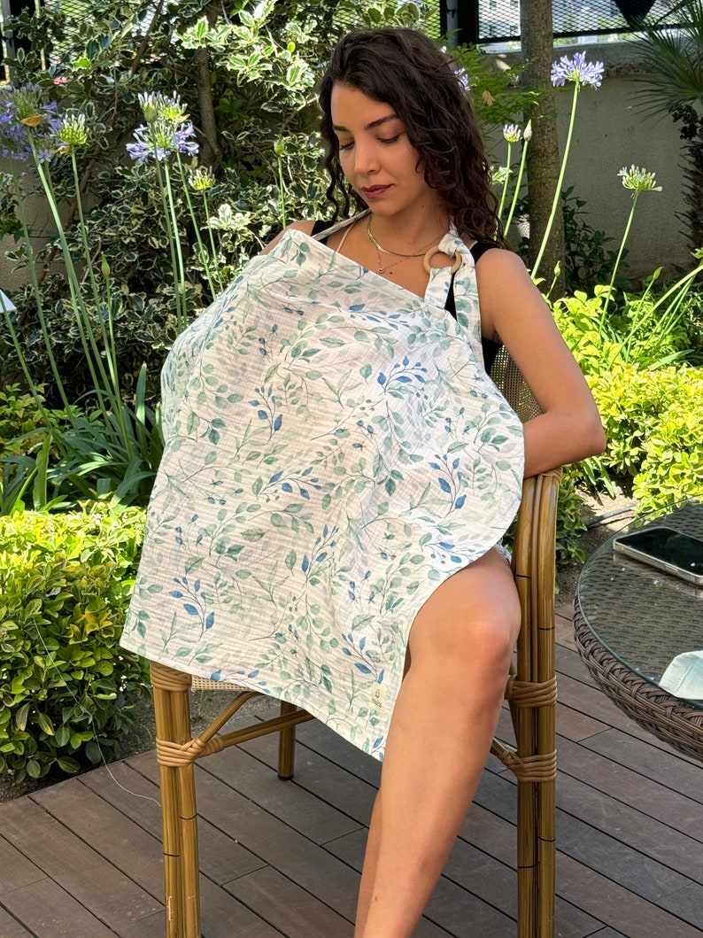 Puede incluir: Una cubierta de lactancia blanca con un patr&oacute;n de hojas verdes y azules. La cubierta tiene un anillo de madera en la parte superior para una f&aacute;cil apertura y cierre.