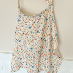 Puede incluir: Una envoltura de beb&eacute; de muselina blanca con un estampado floral en tonos rosa, azul, amarillo y verde. La envoltura est&aacute; colgada en un estante de madera con pomos de colores.