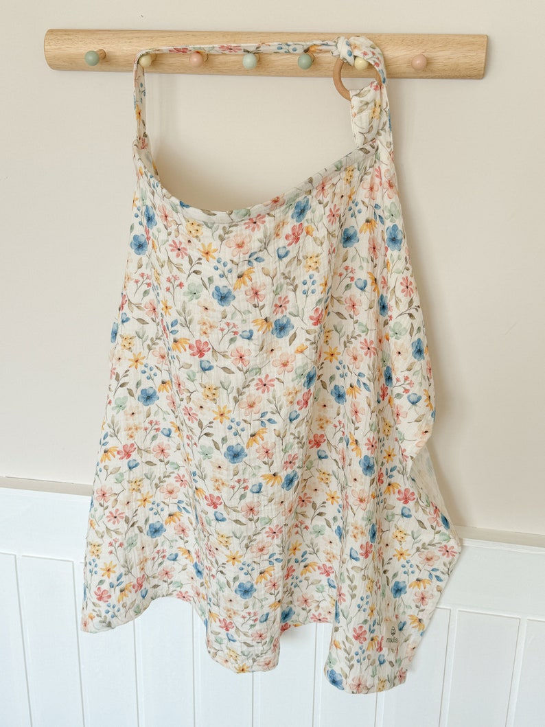 Puede incluir: Una envoltura de beb&eacute; de muselina blanca con un estampado floral en tonos rosa, azul, amarillo y verde. La envoltura est&aacute; colgada en un estante de madera con pomos de colores.