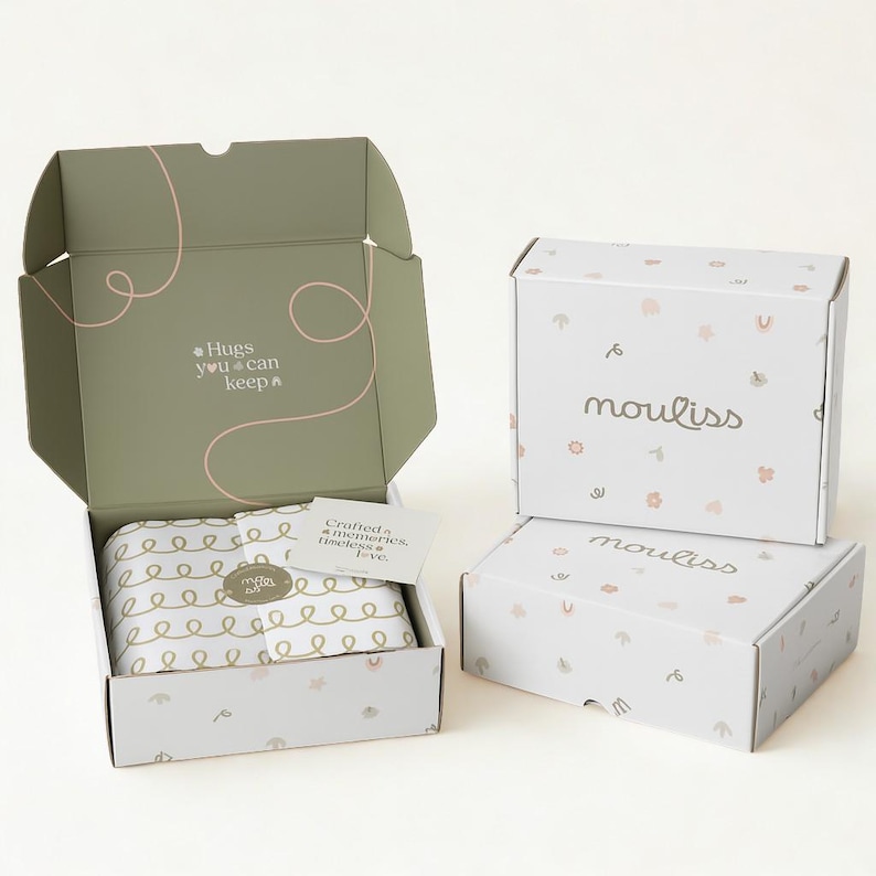May include: Three rectangular cardboard shipping boxes with white exteriors featuring small pastel icons and the word mouliss. One box is open, revealing a sage green interior with the text Hugs you can keep and a card reading Crafted memories, timeless love.