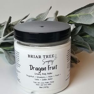 May include: A clear jar of Briar Tree Soapery Dragon Fruit Creamy Body Butter. The label lists ingredients and scents: Dragonfruit, Strawberry, Melon, Peach, Vanilla, and Golden Amber. The jar has a black lid and contains 8 oz (227g) of body butter.