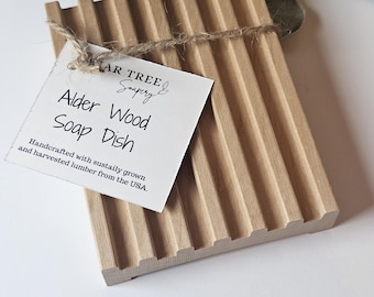 May include: A light brown alder wood soap dish with a grooved design. A tag tied with twine reads "Alder Wood Soap Dish" and mentions sustainably grown lumber from the USA. The soap dish is a natural wood color.