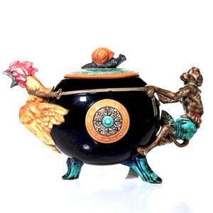 Könnte beinhalten: Eine skurrile Keramik-Teekanne mit schwarzem Korpus, einem Hahnausguss und einem Affengriff. Der Deckel ist mit einer Schneckenfigur verziert. Die Teekanne hat ein kreisförmiges Design an der Seite und steht auf drei türkisfarbenen Füßen.