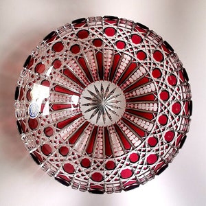May include: A round, deep crystal bowl featuring a deep red and clear glass design with intricate geometric cut patterns. The centre displays a starburst motif, surrounded by radiating panels and a small blue and gold Bohemia sticker on the rim.