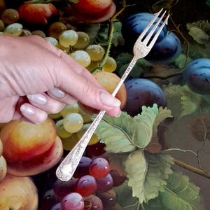 Puede incluir: Un tenedor de metal plateado con tres dientes y un mango grabado y ornamentado con intrincados patrones de volutas. El cuello del tenedor presenta un pequeño grabado detallado, floral o tipo escudo, cerca de la base de los dientes.