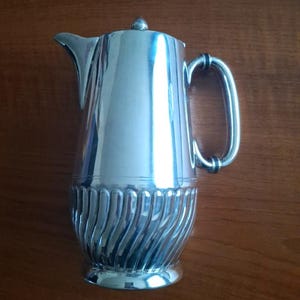 May include: A silver-colored pitcher with a curved spout and a handle. The bottom portion features a ribbed design. The pitcher has a lid with a small knob on top. The background is a wooden surface.